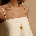 Gold-toned pendant necklace with rectangular charm and decorative leaf detail, worn with beaded choker-style necklace.