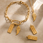 Gold-plated charms including angel wings, numbered rectangular pendants, and a chain bracelet on beige fabric.