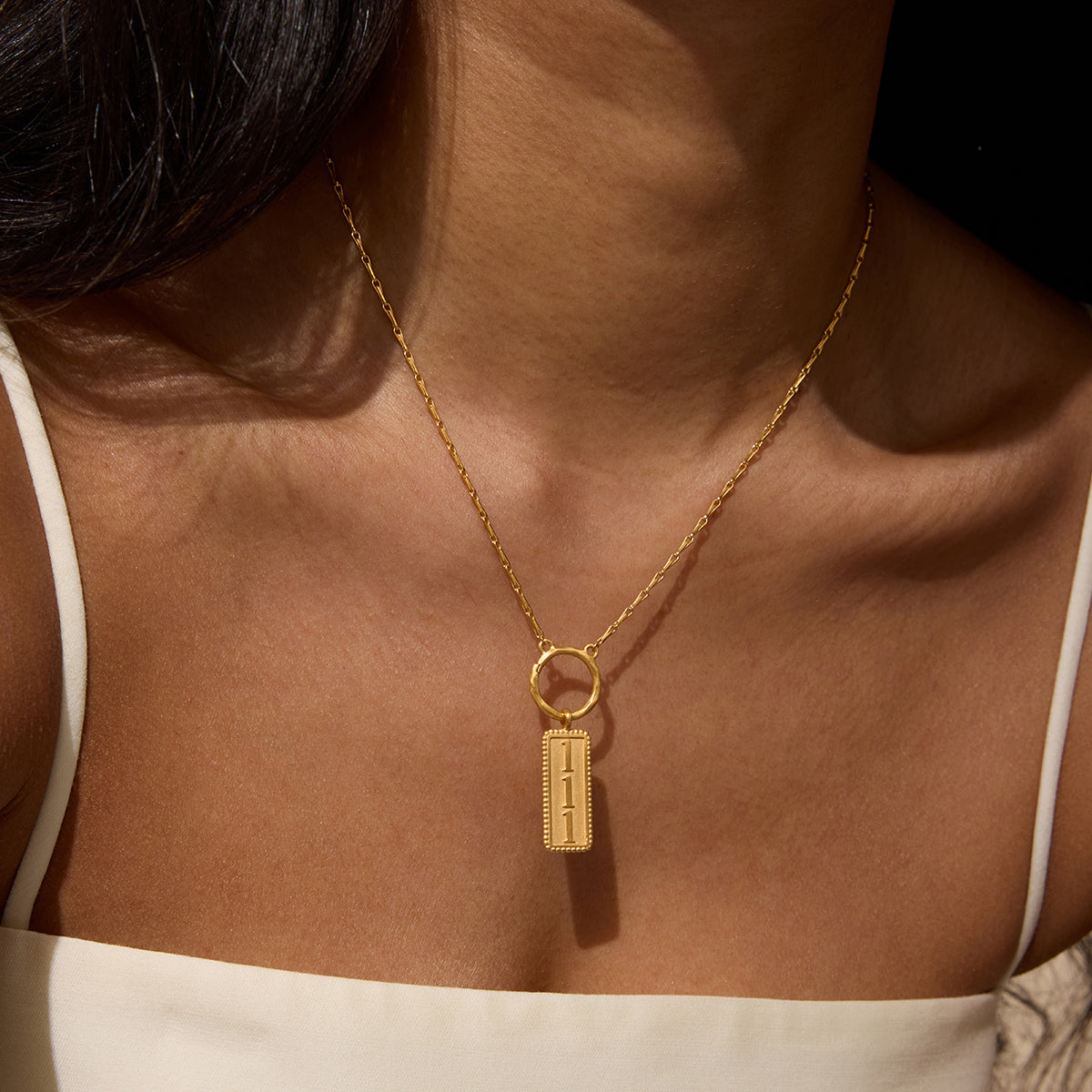 Gold-toned rectangular pendant with "111" engraved on face, hanging from delicate chain worn as necklace.