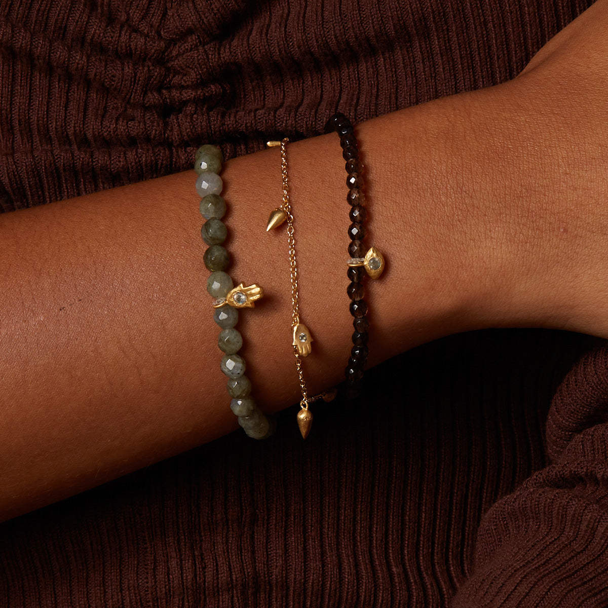 Three beaded bracelets stacked on wrist: gray faceted stone beads, gold chain with pendant, and black beads with gold accents