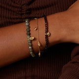 Three beaded bracelets stacked on wrist: gray faceted stone beads, gold chain with pendant, and black beads with gold accents