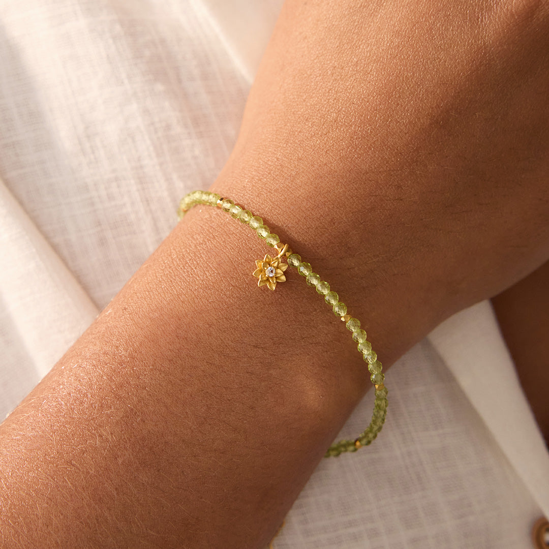 Person wearing the Peridot Lotus August Birthstone Bracelet with vibrant green peridot beads and a gold-plated lotus flower pendant. This piece of spiritual jewelry is crafted for meditation and serves as a protective talisman, symbolizing harmony and new beginnings. A beautiful representation of zodiac energies, this bracelet is ideal for those who engage in yoga and seek meaningful connection.