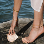 Gold-toned beaded anklets worn on person standing on rock near water.
