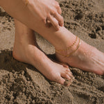 Two gold-toned chain anklets worn on a person's ankle at the beach.