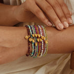 Stack of beaded bracelets with purple, white, green, and gold beads and gold lotus flower charms.