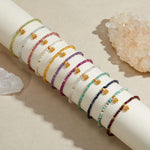 Multiple beaded bracelets in various colors with gold lotus charms, displayed on a white cylindrical stand next to a rose qua