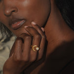 Gold-toned ring with pale moonstone stone worn on ring finger of person with deep skin tone.