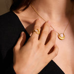 Gold-toned ring with pale stone center worn on hand, paired with matching pendant necklace.