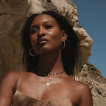 Timeless Beauty Dot Hoop Earrings showcased on a woman with radiant skin, exuding confidence against a dramatic backdrop. This elegant piece of spiritual jewelry symbolizes truth, perfect for meditation and manifestation. Crafted with 18kt gold plate, these earrings are ideal for yoga enthusiasts seeking meaningful adornments that reflect protection and connection to the sun and moon.