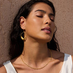 Gold petal-shaped drop earrings with curved design worn on person against textured beige background.