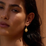 Gold teardrop earring with textured finish worn on earlobe against warm-toned background.