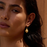Gold teardrop earring with textured finish worn on earlobe against warm-toned background.