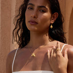 Gold drop earrings with iridescent opal stone centers, worn by woman with dark hair against warm beige background.