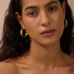 Gold-toned hammered hoop earrings worn on model with long dark hair against neutral background.