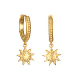 Pair of gold huggie earrings with textured oval hoops and dangling sun-shaped charms with hammered texture.