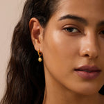 Gold sun-shaped huggie earrings with pointed rays hanging from the earlobe on a person with long dark hair.