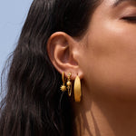 Gold huggie earrings with sun charm pendants worn on ear lobe against blue background.