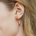Gold triangle hoop earring worn on earlobe, elongated V-shaped design with polished finish.