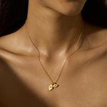 A fine gold chain necklace with two small pendants, a hamsa hand and an eye charm, rests on a bare collarbone.