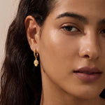 Gold-toned huggie hoop earrings with hamsa hand charms hanging from the hoops, worn on a model's ears.