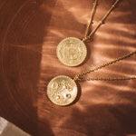 Two gold pendant necklaces with circular medallions featuring ornate floral and mandala designs on brown fabric.