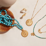 Turquoise beaded mala necklace with gold sun medallion charm draped over wooden bowl, displayed with coordinating gold and ge