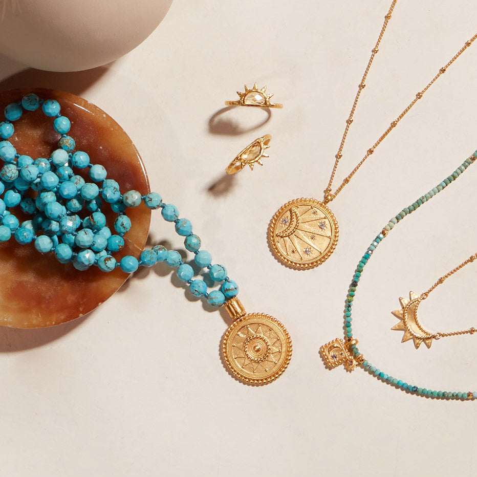 Turquoise beaded mala necklace with gold sun medallion charm draped over wooden bowl, displayed with coordinating gold and ge