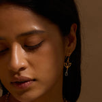 Close-up of a woman with dark hair looking downward, wearing a gold lotus-shaped drop earring with a pale blue teardrop stone.