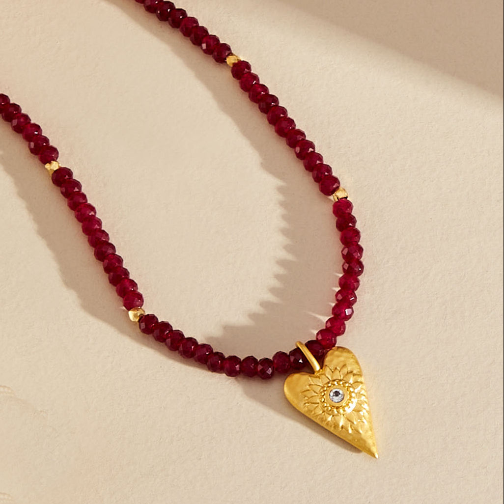A beaded necklace featuring deep red faceted beads and a textured gold heart-shaped pendant with a small blue stone, displayed on a warm beige background.