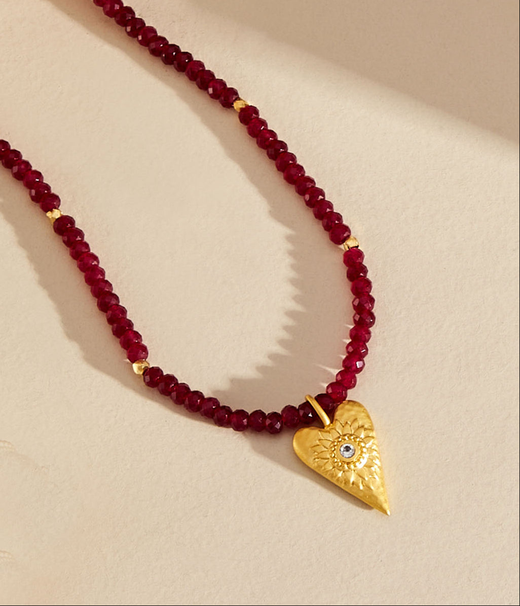 A beaded necklace featuring deep red faceted beads and a textured gold heart-shaped pendant with a small blue stone, displayed on a warm beige background.