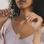 Beaded mala necklace with turquoise and green stones, gold circular pendant, worn as long strand.