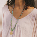 Beaded mala necklace with turquoise, lavender, and green stones, gold circular pendant, worn over light pink v-neck shirt.