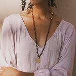 Long beaded mala necklace with red, black, and natural stone beads, gold circular pendant, worn over lavender top.