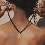 Beaded mala necklace with dark red and black stones and gold medallion pendant worn around person's neck.