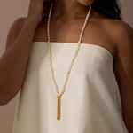 Long beaded necklace with white mother of pearl round beads and gold tassel pendant worn over cream-colored garment.