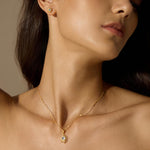A woman with long dark hair wears a gold starburst pendant necklace with a pale stone and a matching stud earring.