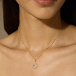 A woman wears a fine gold chain necklace with a sunburst pendant holding a round clear gemstone at its center.