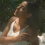 Gold lariat necklace with lotus pendant worn long on person in white tank top against green foliage background.