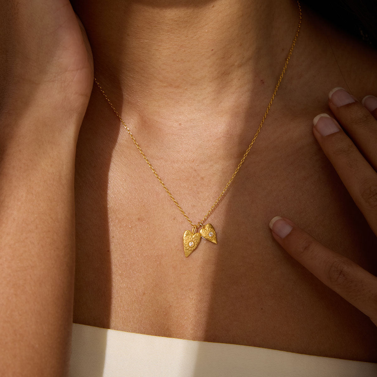 A gold chain necklace with two small textured heart pendants, each set with a tiny clear stone, rests on a person's collarbone.