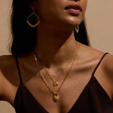 A woman in a dark camisole wears layered gold necklaces with heart pendants and large teardrop-shaped gold earrings.
