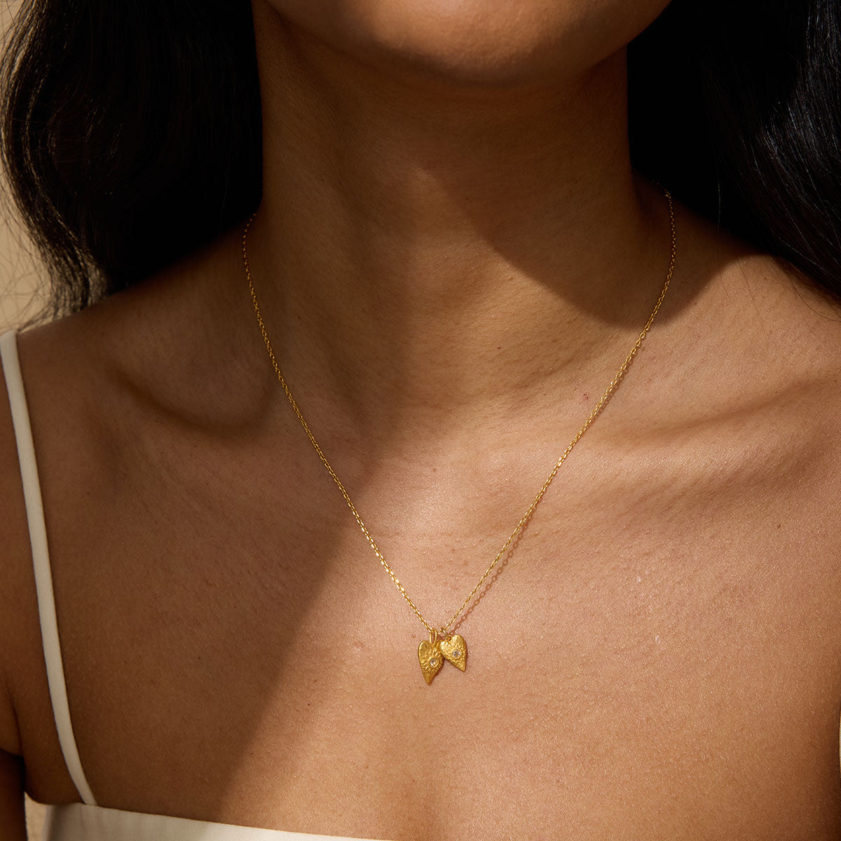A woman wearing a thin gold chain necklace with two small heart-shaped pendants resting at her collarbone, dressed in a white strap top.