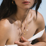 Gold chain necklace with small white opal and shell charms, worn layered on a person's chest.