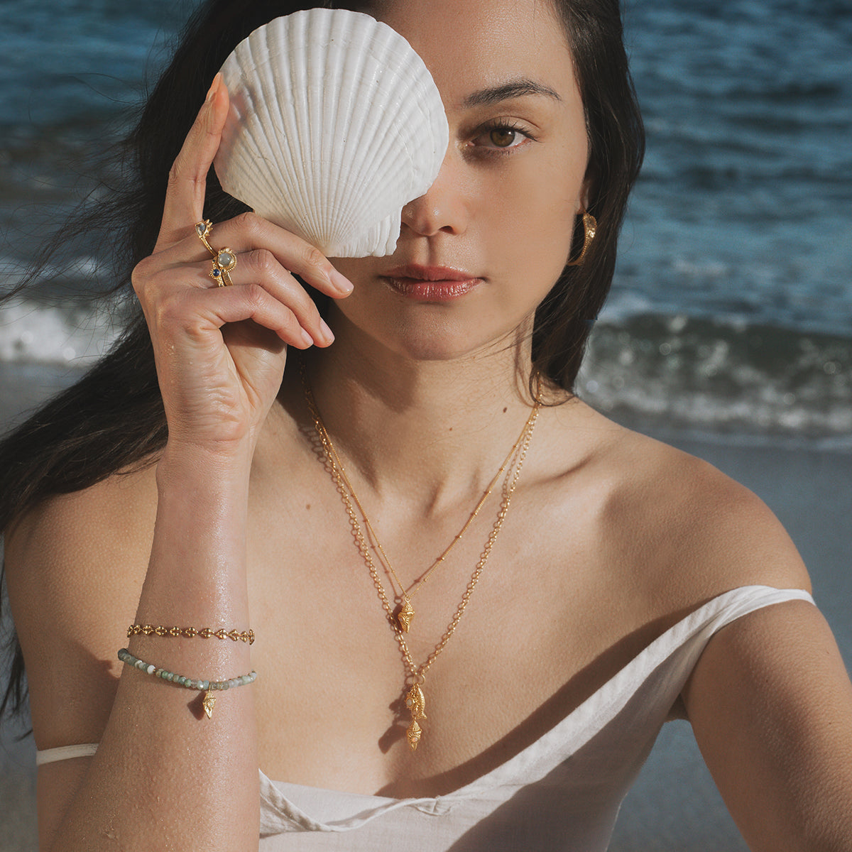 Gold-toned necklace with small pendant worn on delicate chain, displayed on model at beach.