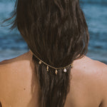 Gold chain necklace with shell and pearl charms worn around braided dark hair at the beach.