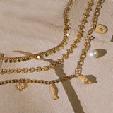 Gold-toned layered chains with pearl, fish, sun, and shell charms on sand background.