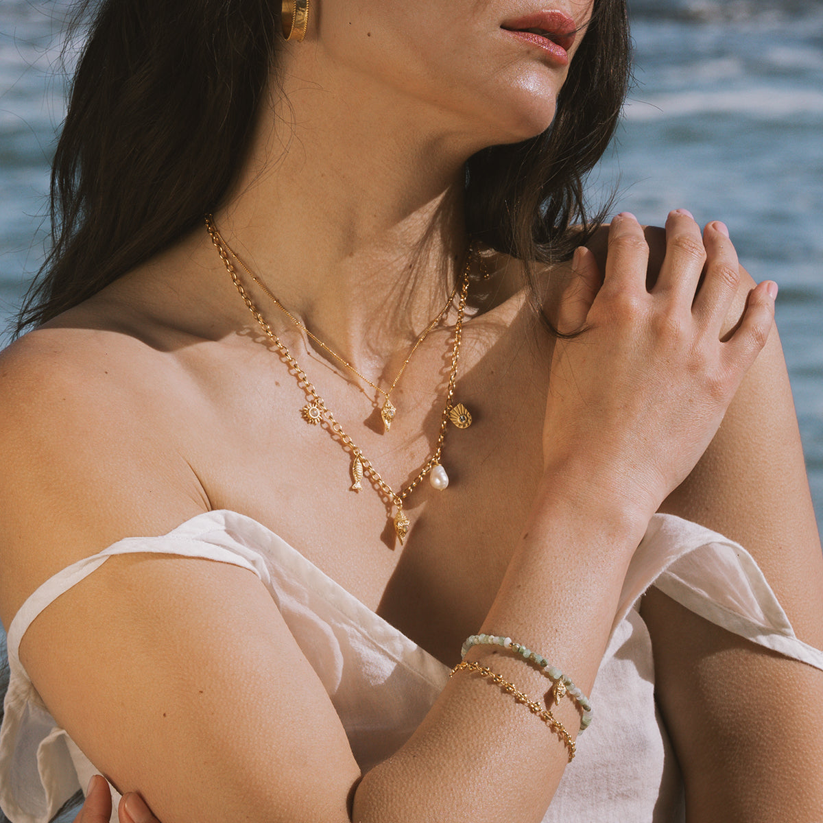 Gold-toned layered necklaces with shell and pearl charms worn on person near ocean water.