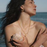 Gold chain necklace with pearl and shell-shaped charm pendant worn by woman at ocean.
