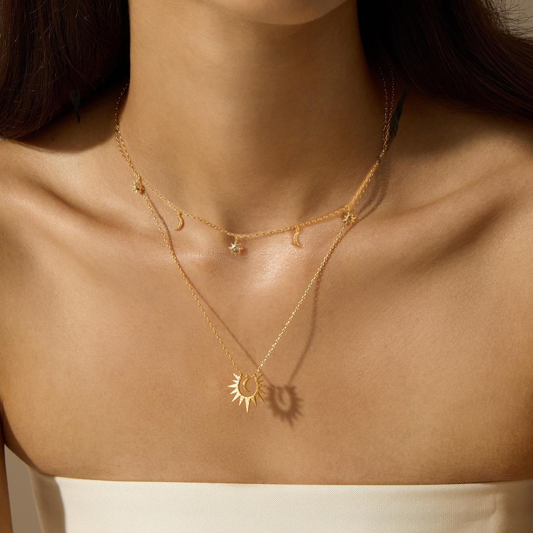 A woman wears two layered gold necklaces, one with small moon and star charms and one with a sunburst pendant.