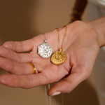 A hand holding two pendants, the Mother Moon Silver Locket Necklace displayed alongside a golden version. Both symbolize spiritual growth and serve as talismans for protection, ideal for yoga and meditation enthusiasts seeking meaningful adornments.