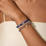 Three beaded bracelets stacked on wrist: dark blue sodalite, light rose quartz, and gray gemstone strands with gold moon char
